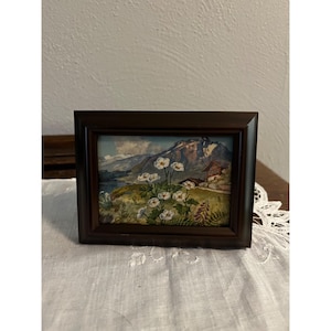 May include: A framed landscape painting featuring a mountain range, a lake, and a small cabin. White flowers are in the foreground. The frame is dark brown. The painting is displayed on a white lace tablecloth.