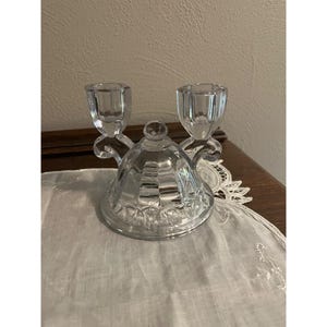 May include: A clear glass candle holder with two candle cups. The base has a decorative, faceted design. The candle holder is sitting on a white, embroidered cloth.
