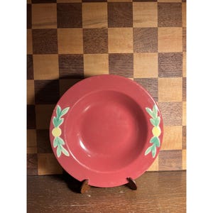 May include: A red ceramic plate with a shallow bowl shape. The plate has a decorative design of green leaves and yellow circles. The plate is displayed on a wooden stand against a checkered background.