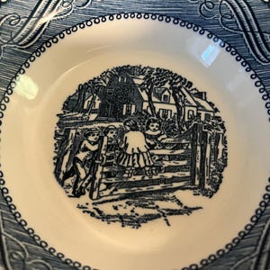 May include: A blue and white ceramic plate with a black and white illustration of children playing by a fence in front of a house. The plate has a blue and white border with a repeating pattern.