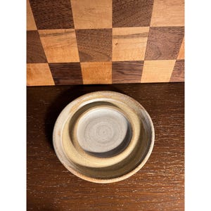 May include: A handmade, ceramic, beige-colored dish with a circular design. The dish has a raised, concentric inner circle, creating a small, recessed area. The outer rim is a lighter shade of beige. The dish is on a dark brown wooden surface.