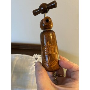 May include: A wooden corkscrew with a carved flower design. The corkscrew has a dark brown finish and a metal screw. The handle is cylindrical with a decorative flower carved into the wood.