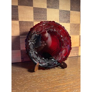 May include: A ruby red glass plate with a decorative, embossed pattern. The plate is round with a scalloped edge and is displayed on a small wooden stand. The background features a wooden checkerboard pattern.