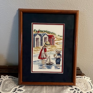 May include: Framed cross-stitch artwork depicting two children playing near the beach. The artwork features a sailboat, beach huts, and children in vintage clothing. The frame is a dark brown color with a navy blue matting.