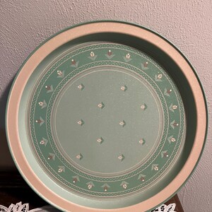 May include: Round serving tray with a light green center and a pinkish-beige border. The center features a floral pattern in white, green, and brown. The tray is likely made of metal or a similar material and is designed for serving food or drinks.