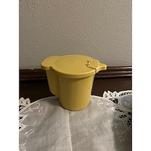 May include: A yellow plastic pitcher with a matching lid. The pitcher has a handle and a spout. The lid has a small tab for opening and closing. The pitcher is sitting on a white lace doily.