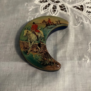 Pode incluir: Uma caixa de bugigangas vintage em forma de crescente com uma cena de caça. A caixa apresenta uma ilustração pintada de cavaleiros a cavalo, um cão e uma paisagem. A caixa tem uma borda azul e é colocada sobre um fundo branco.