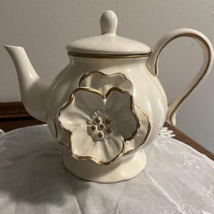 May include: Cream-colored ceramic teapot with a large, three-dimensional flower design on the front. The flower and the edges of the teapot are accented with gold trim. The teapot has a lid and a curved handle.