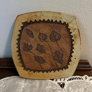May include: Square ceramic plate with a brown leaf design. The plate has a tan border with a dark brown zigzag pattern. The center of the plate is a darker brown with six brown leaves.