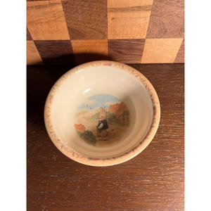 May include: A small, off-white ceramic bowl with a decorative rim. The bowl features a charming illustration of a rabbit in a blue jacket standing in a field. The rim is decorated with a floral pattern.