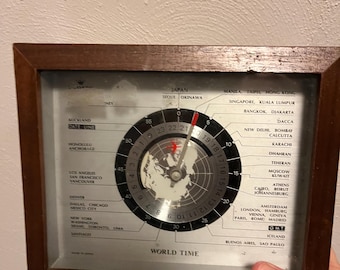 May include: A vintage world time clock with a dark wooden frame. The clock face features a map of the world and displays various international cities. The hands indicate the current time. The text "WORLD TIME" is visible.