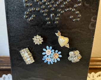 May include: A collection of vintage brooches and pins displayed on a black metal container. The brooches include a rhinestone flower, a heart with a pearl, a rectangular design, a cluster of blue and pink flowers, and a round rhinestone brooch.