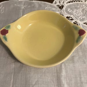 May include: A small, yellow ceramic dish with two handles. The dish has a rounded shape with a slightly raised rim. Each handle is decorated with a red rose and green leaves. The dish is likely used for serving small portions of food.