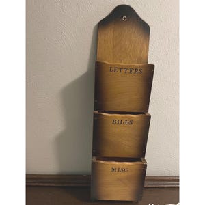 May include: A wooden wall-mounted organizer with three compartments labeled "LETTERS", "BILLS", and "MISC". The organizer is made of stained wood with a dark top and a hole for hanging. The compartments are stacked vertically.