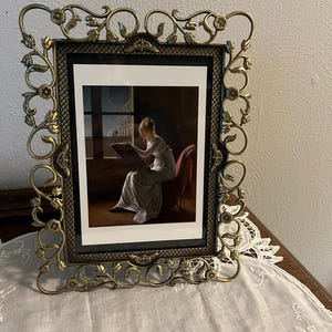 May include: Ornate brass picture frame with a floral and scroll design. The frame surrounds a print of a woman reading by a window. The frame is displayed on a wooden surface covered with a white lace cloth.