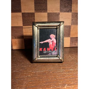 May include: A rectangular metal frame with an ornate, embossed scrollwork pattern featuring a centered image of a person in a red swimsuit. The text KAMEL Cigarettes is printed in red and white lettering at the bottom of the central image.
