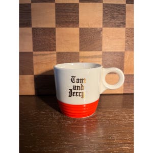 May include: A white ceramic mug with a red base and a rounded handle. The mug features the text "Tom and Jerry" in gold lettering. The mug is sitting on a wooden surface.