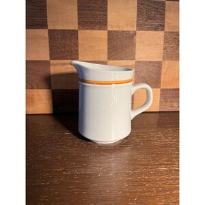 May include: A small, white ceramic creamer with a handle and a spout. The creamer has a horizontal orange stripe near the top. The creamer is sitting on a wooden surface.