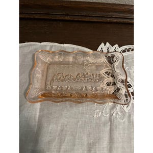 May include: A rectangular, pink glass dish with a scalloped edge. The dish features a detailed relief of the Last Supper. The dish is resting on a white, embroidered cloth.