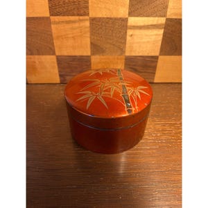 May include: A small, round, red lacquered box with a bamboo design on the lid. The bamboo is depicted in gold and black. The box is sitting on a wooden surface.
