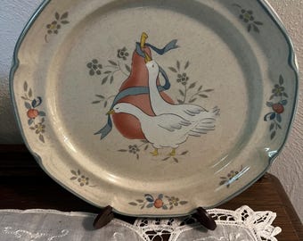May include: A decorative plate with a scalloped edge and a light blue border. The plate features a pear with a blue ribbon and two white geese. Floral and fruit accents are scattered around the rim. The plate is displayed on a wooden stand.