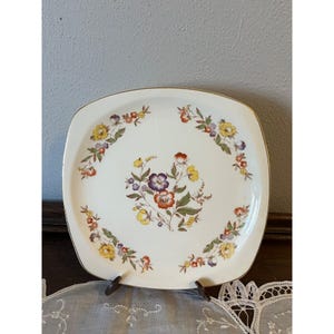 May include: Square, off-white ceramic plate with a gold rim and floral design. The plate features a central cluster of purple, orange, and yellow flowers, surrounded by a floral border. The plate is displayed on a brown stand.