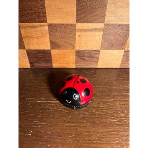 May include: A red and black ladybug-shaped kitchen timer. The timer has a black face with white markings and a dial. The ladybug has black spots on its red shell. The timer is sitting on a wooden surface.