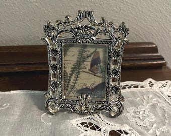 May include: Ornate silver-colored picture frame with a rectangular opening. The frame features intricate floral and scrollwork designs. The picture inside shows a butterfly and botanical print. The frame is resting on a white lace doily.
