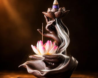 May include: A dark brown ceramic incense burner shaped like two hands cupping a pink and white lotus flower. A lit incense cone sits atop the upper lotus, releasing white smoke that cascades downward over the fingers into the lower lotus base.