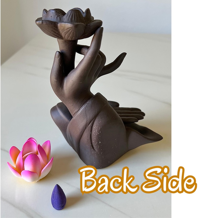 May include: A dark brown ceramic incense burner shaped like a hand holding a lotus flower, accompanied by a small pink and yellow artificial lotus flower and a purple cone incense. The text "Back Side" is printed in orange on the lower right.