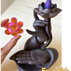 May include: A dark brown ceramic incense burner shaped like a hand holding a lotus flower, with a purple cone incense placed on top. A separate, smaller pink and orange lotus flower accessory is held nearby. The text Front Side is visible.