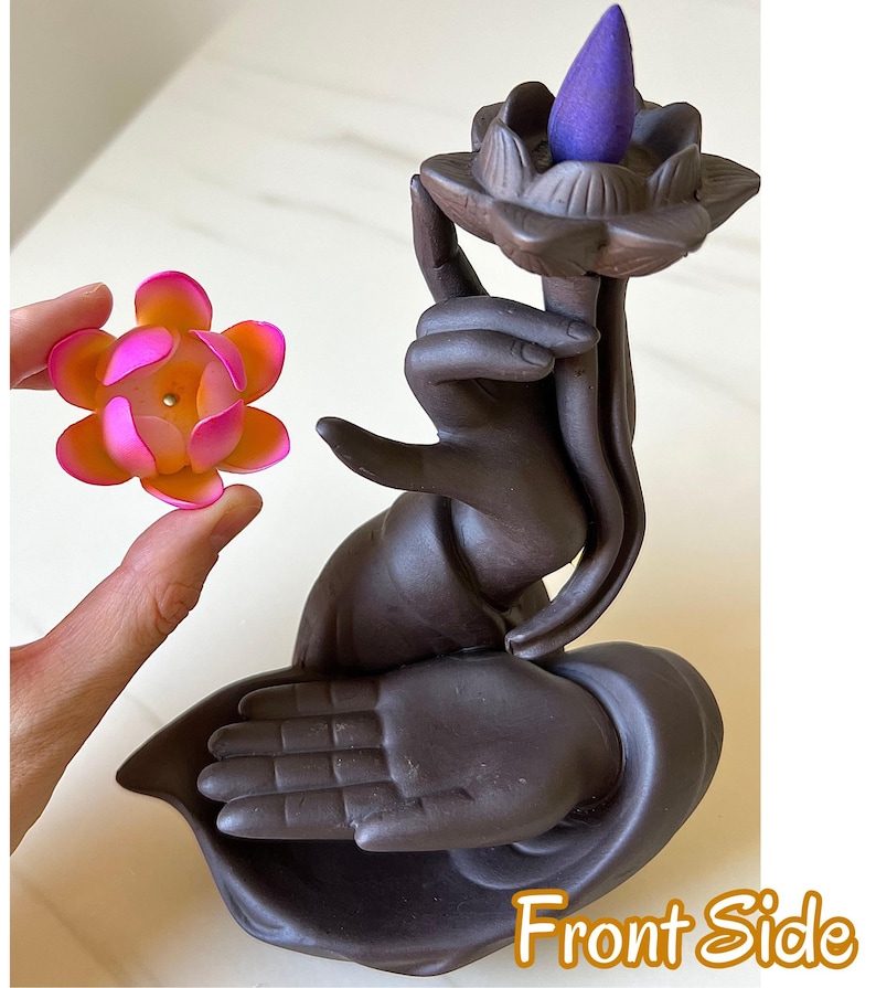 May include: A dark brown ceramic incense burner shaped like a hand holding a lotus flower, with a purple cone incense placed on top. A separate, smaller pink and orange lotus flower accessory is held nearby. The text Front Side is visible.