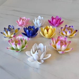 Puede incluir: Una colección de flores de loto artificiales en varios colores, incluyendo rosa, blanco, amarillo, azul y morado. Las flores están dispuestas sobre una superficie de mármol blanco, mostrando sus detalladas estructuras de pétalos.