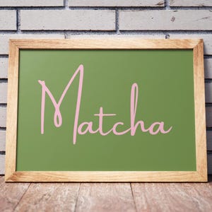 Könnte beinhalten: Ein gerahmtes Schild mit grünem Hintergrund und dem Wort "Matcha" in rosa Schrift. Der Rahmen besteht aus hellbraunem Holz und steht vor einer weißen Backsteinmauer und einer Holzoberfläche.