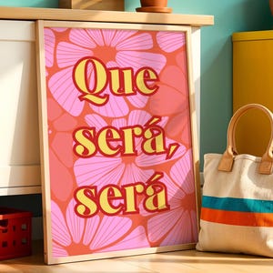 Könnte beinhalten: Ein gerahmter Druck mit den Worten "Que será, será" in gelber Schrift auf einem rosa Blumengrund. Eine Canvas-Tragetasche mit blauen und orangefarbenen Streifen und einem Holzgriff steht neben dem Druck.