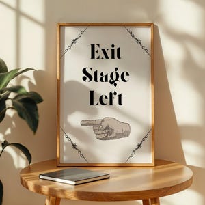 May include: A framed print with the words "Exit Stage Left" in black, with a hand pointing left. The print is in a wooden frame and sits on a round wooden table. A closed notebook is on the table.