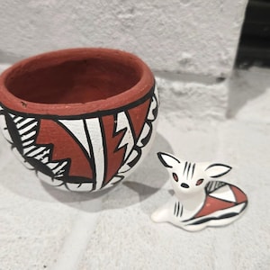 May include: A round terracotta bowl with a wide opening and a hand-painted geometric pattern in black, white, and reddish-brown. Beside the bowl sits a small, matching ceramic figurine of a reclining deer with similar black, white, and brown details.