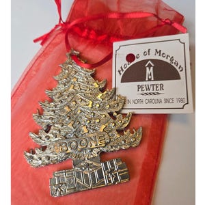 May include: A silver-colored pewter Christmas tree ornament with the word "BOONE" on it, displayed in a red organza gift bag. The ornament has a detailed tree design and is from "House of Morgan, Pewter, in North Carolina since 1980."