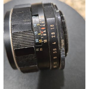 以下が含まれることがあります： 複数のリングとマーキングが付いた黒いカメラレンズ。レンズにはフィートとメートルの距離表示があり、絞り設定は1.8から16まであります。レンズには「AUTO」設定もあります。