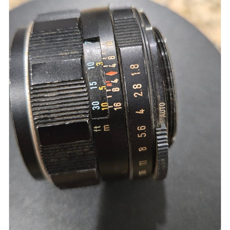 以下が含まれることがあります： 複数のリングとマーキングが付いた黒いカメラレンズ。レンズにはフィートとメートルの距離表示があり、絞り設定は1.8から16まであります。レンズには「AUTO」設定もあります。