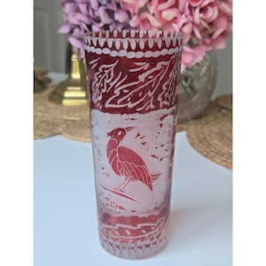 May include: A tall, cylindrical glass vase with a deep red hue. The vase features intricate etched designs, including a bird and ornate patterns. The rim and base have detailed decorative elements. The vase is likely used for displaying flowers.