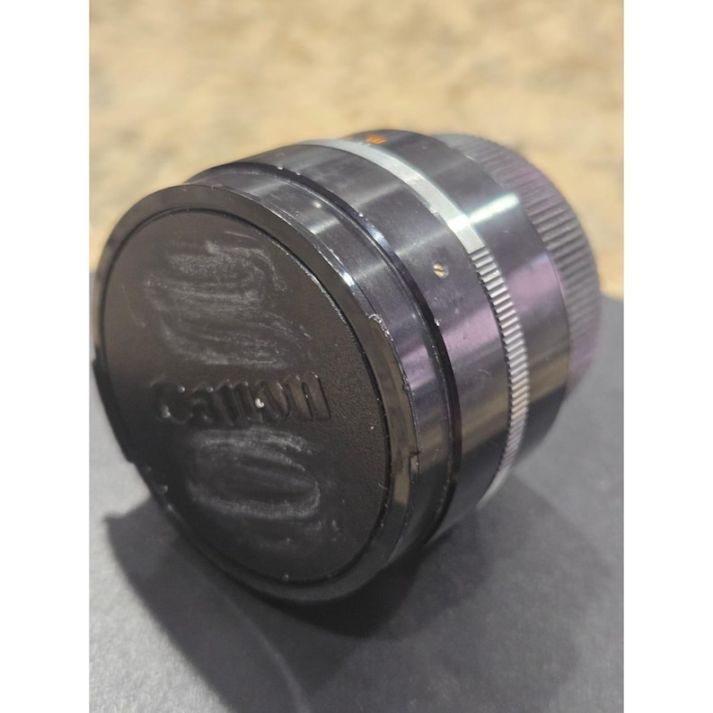 May include: A Canon camera lens with a black lens cap. The lens is silver and black with ridged edges for grip. The Canon logo is visible on the lens cap. The lens is designed for photography.