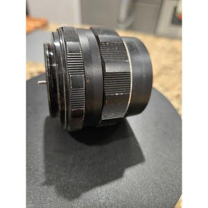 以下が含まれることがあります： 黒いカメラレンズアダプター。エッジには溝があり、銀色のピンが付いています。アダプターは円筒形で、金属製に見えます。レンズアダプターは、さまざまなカメラコンポーネントを接続するように設計されています。