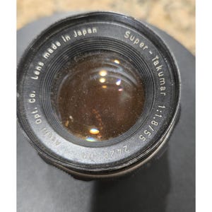 以下が含まれることがあります： 黒とシルバーのカメラレンズのクローズアップ。レンズには「Super-Takumar 1:1.8/55」と「Lens made in Japan」のマークがあります。レンズは反射面を持っています。