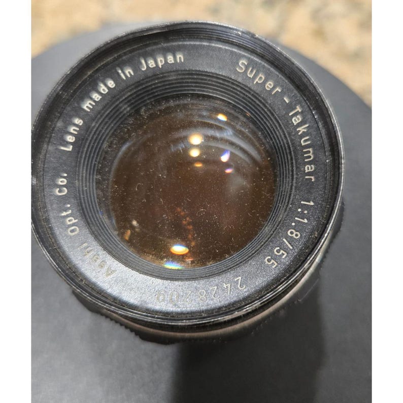 以下が含まれることがあります： 黒とシルバーのカメラレンズのクローズアップ。レンズには「Super-Takumar 1:1.8/55」と「Lens made in Japan」のマークがあります。レンズは反射面を持っています。