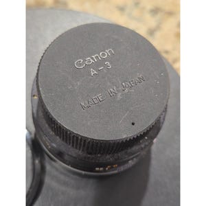 May include: A close-up of a black Canon A-3 lens cap. The words "Canon A-3" are printed in white, with "MADE IN JAPAN" below. The cap is round with a ridged edge.