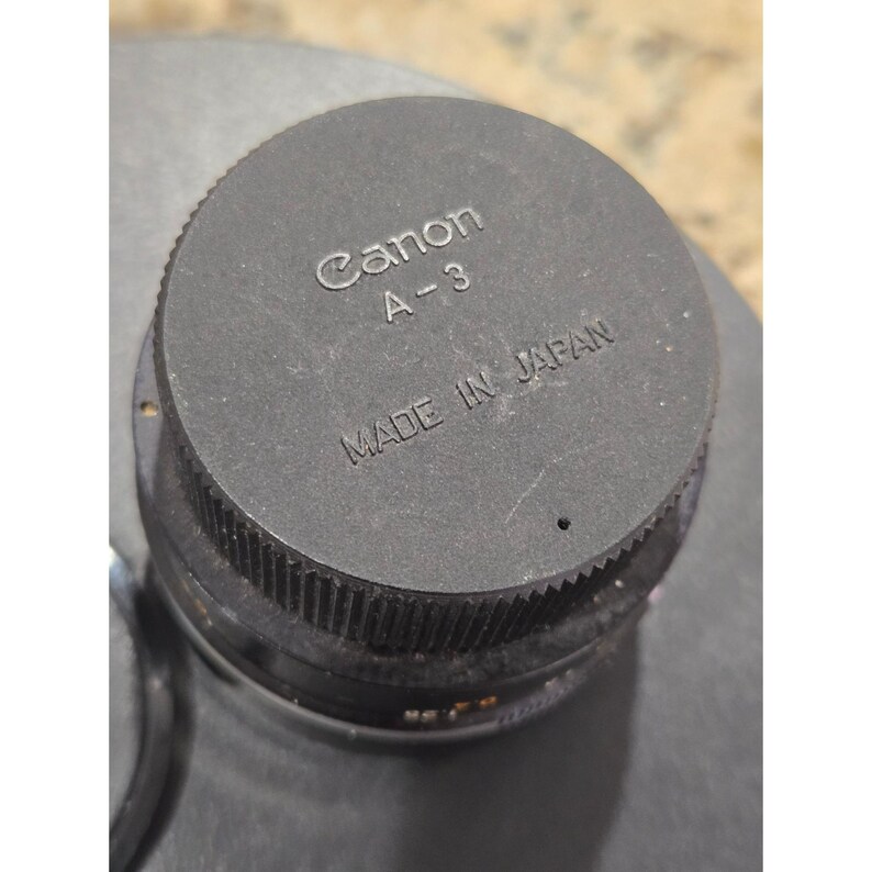 May include: A close-up of a black Canon A-3 lens cap. The words "Canon A-3" are printed in white, with "MADE IN JAPAN" below. The cap is round with a ridged edge.
