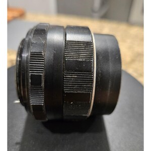 以下が含まれることがあります： 黒いカメラレンズで、溝とテクスチャ加工が施されています。レンズは円筒形で、一連のリングと白いバンドが付いています。レンズは金属製で、写真撮影用に設計されています。