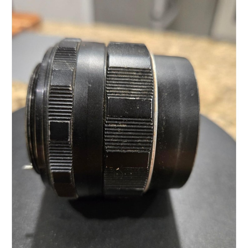 以下が含まれることがあります： 黒いカメラレンズで、溝とテクスチャ加工が施されています。レンズは円筒形で、一連のリングと白いバンドが付いています。レンズは金属製で、写真撮影用に設計されています。