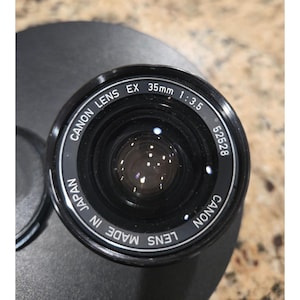 May include: A white ceramic mug with a black handle and the text "But First, Coffee" in a bold, black font. The mug is set against a blurred background, suggesting a cozy setting. The mug appears to be approximately 4 inches tall.